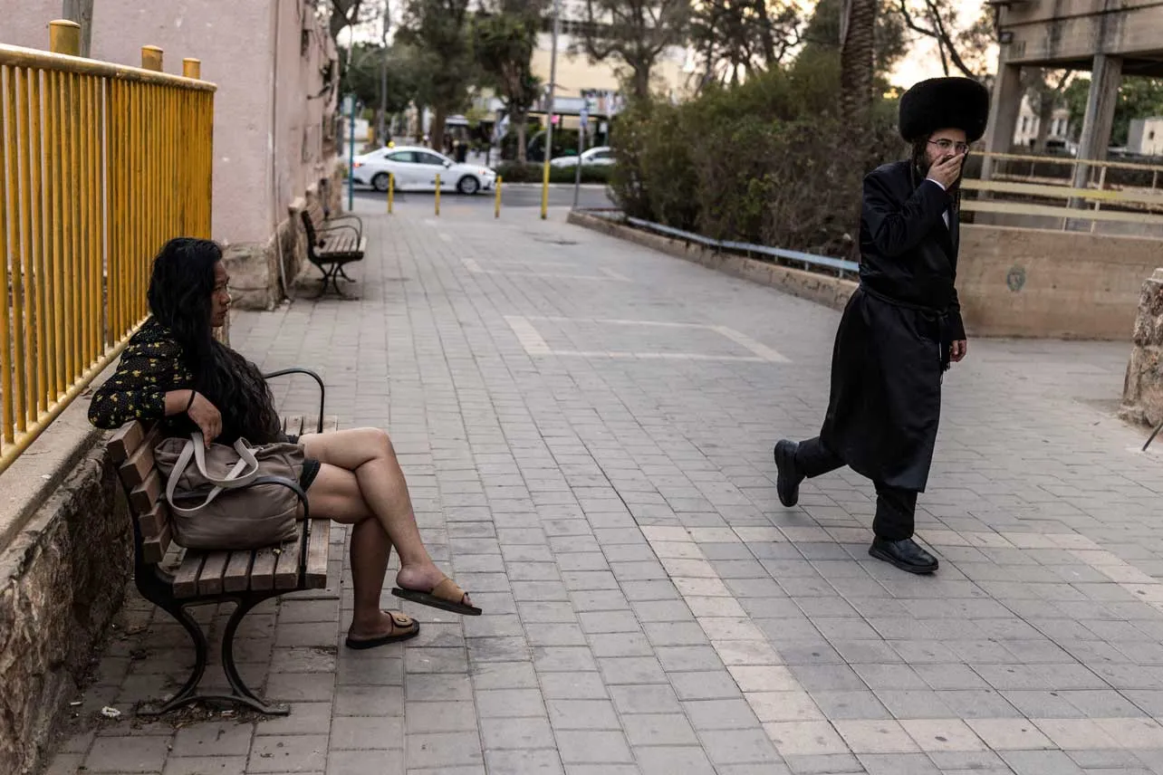 Un judío ultraortodoxo camina por la calle mientras una mujer está sentada en una banca en Israel.