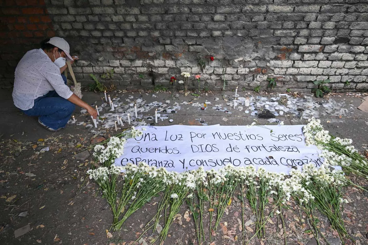 Una mujer enciende velas junto a una manta que dice "Una luz por nuestros seres queridos. Dios dé fortaleza, esperanza y consuelo a cada corazón".